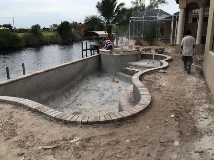 Swimming pool construction beside a canal, featuring curved concrete walls and landscaping work.