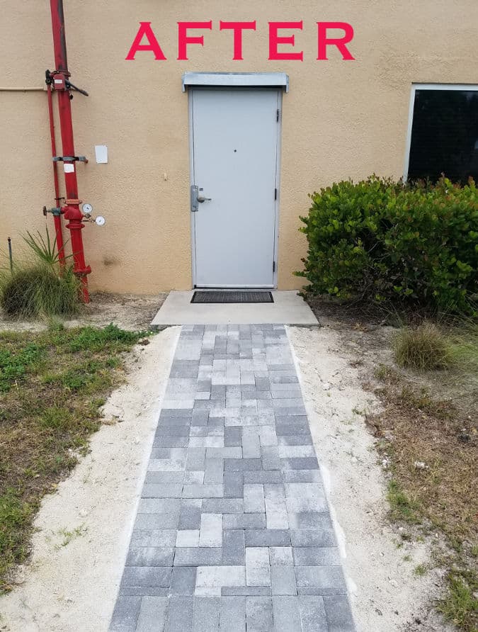 Newly paved pathway leading to a grey door, surrounded by green bushes and grass.