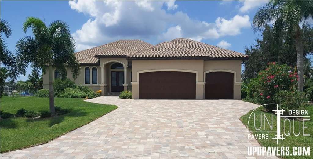 Luxurious home exterior with paver driveway, lush landscaping, and tile roof under blue sky.