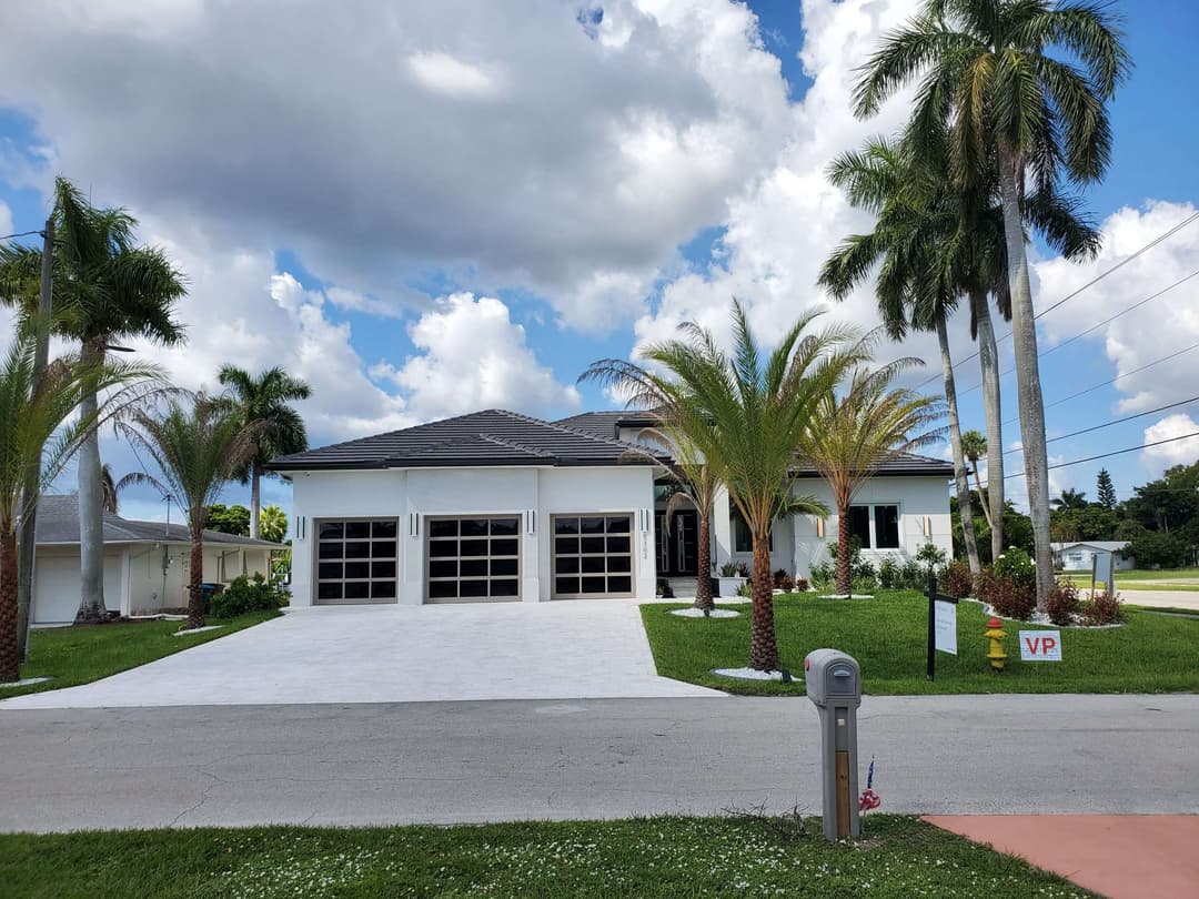 Modern white home with a landscaped yard, palm trees, and a triple garage under a blue sky.
