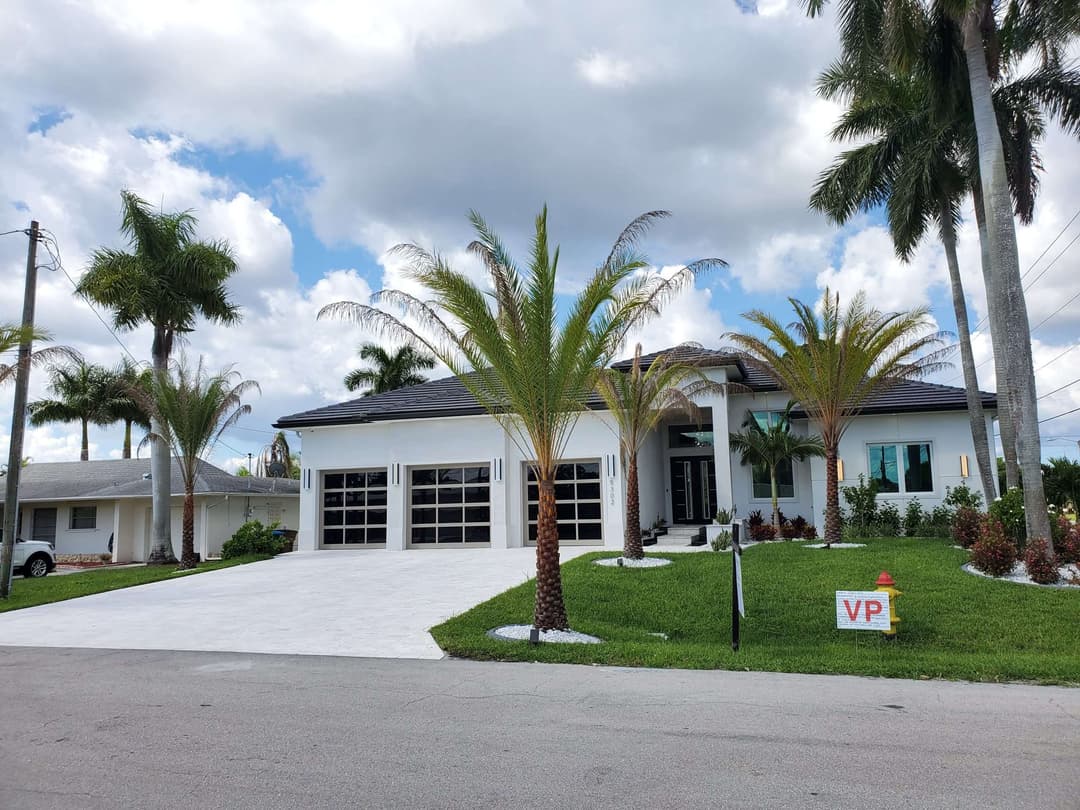 Modern white house with palm trees, large driveway, and vibrant green lawn under a cloudy sky.