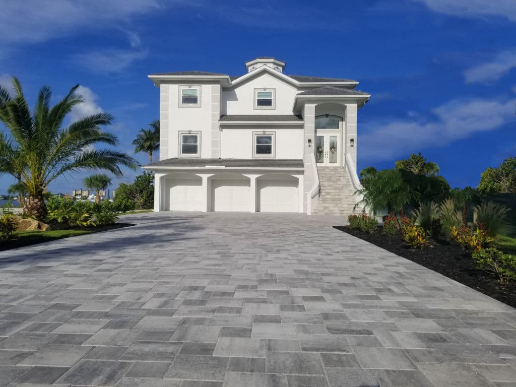 Luxury beach house with a paved driveway and tropical landscaping under a blue sky.