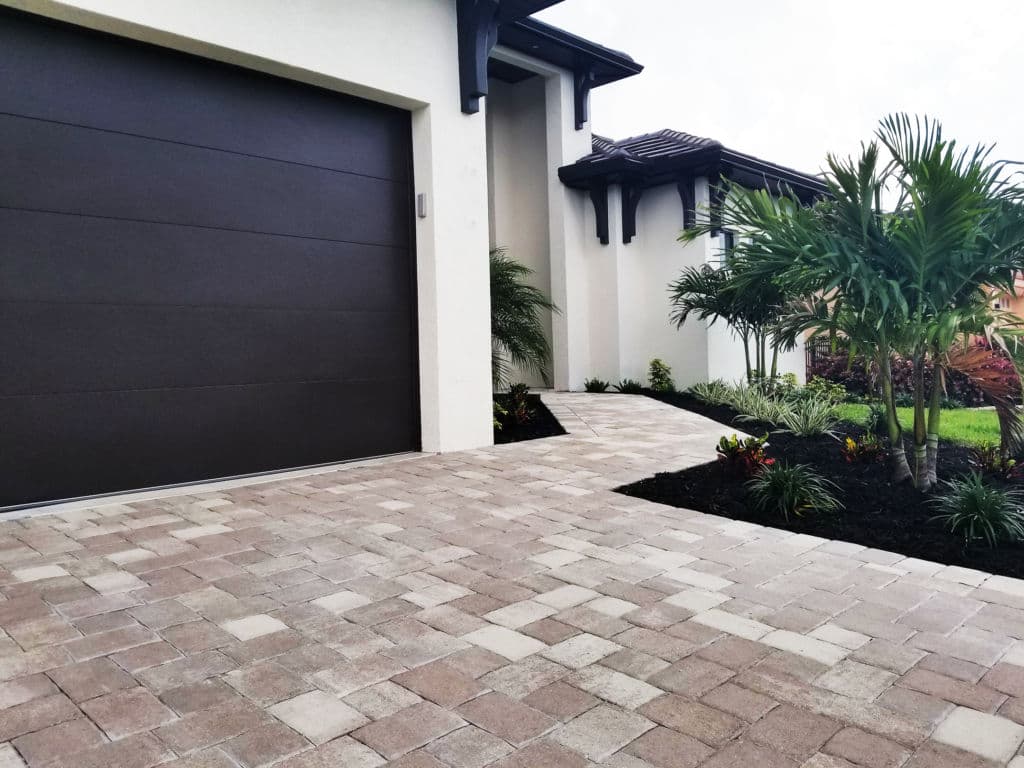 Modern home entrance with paver driveway, lush landscaping, and sleek garage door.