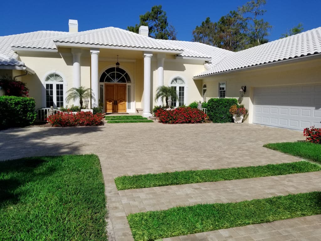 Elegant white house entrance with landscaped gardens and a paved driveway. Bright, sunny day.