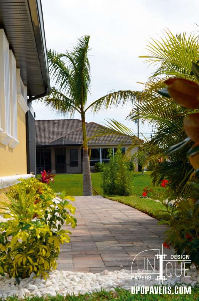 Pathway lined with plants leading to a house with pavers and palm trees in the garden.