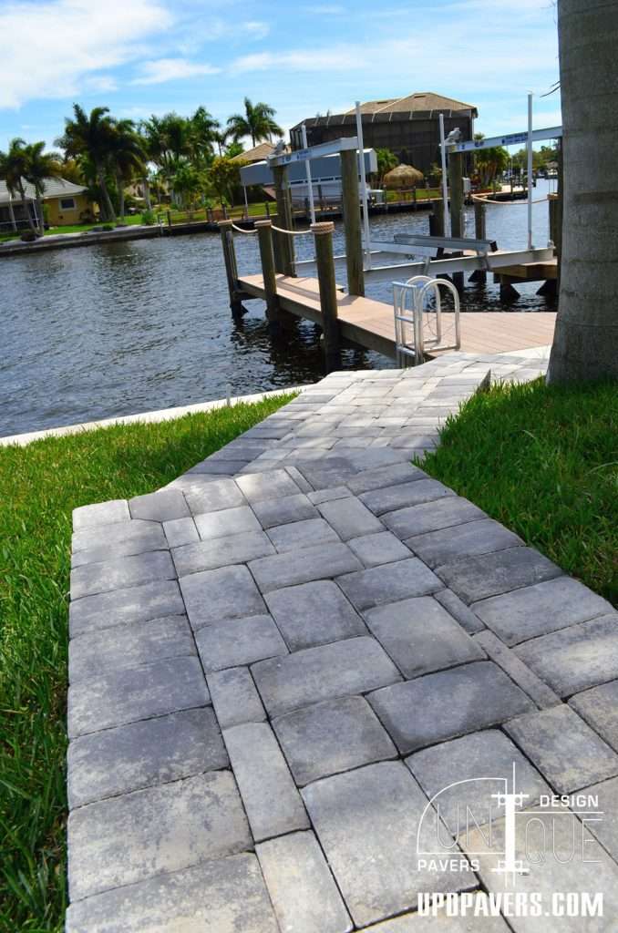 Paver pathway leading to a dock by the water with lush greenery and clear blue sky.