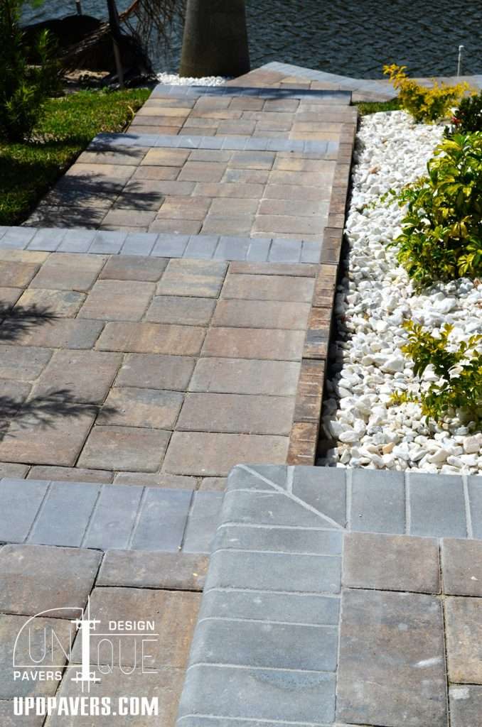 Paver pathway by water with decorative stones and lush greenery, enhancing outdoor aesthetics.