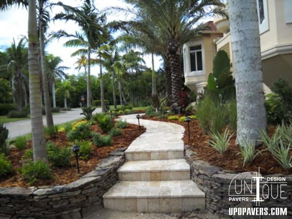 Curved stone pathway surrounded by tropical plants and palm trees near a house.