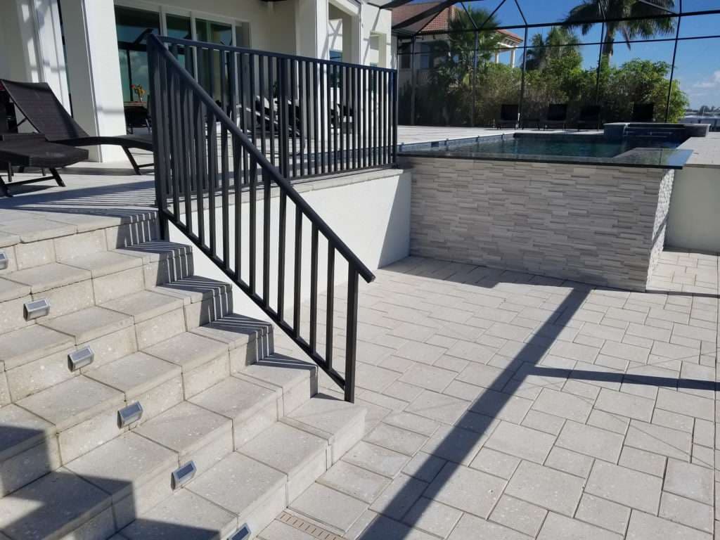 Modern outdoor patio with stairs, railing, and a pool, featuring stone and paver design.