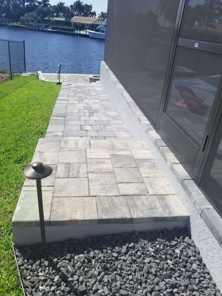Paver walkway by water, leading to a house with landscaping and lighting fixtures.