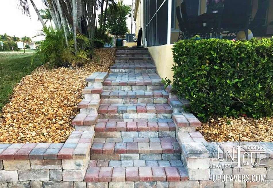 Paver stone pathway with steps leading to a house, surrounded by greenery and landscaping.