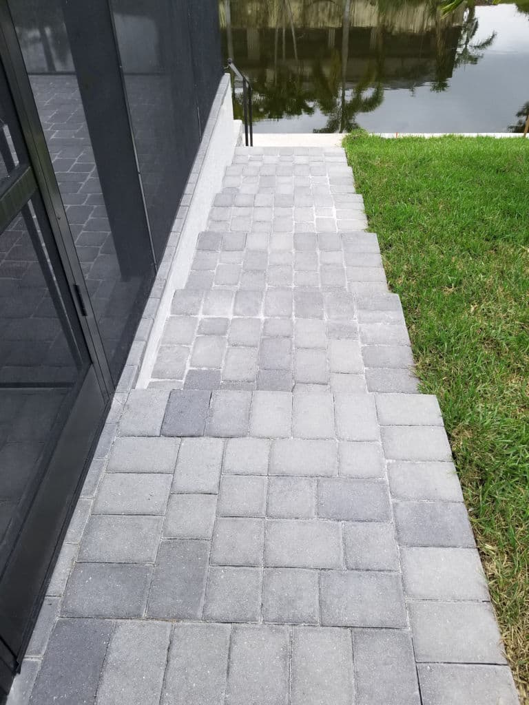 Gray paver stone walkway with steps leading to a waterfront, surrounded by grass.