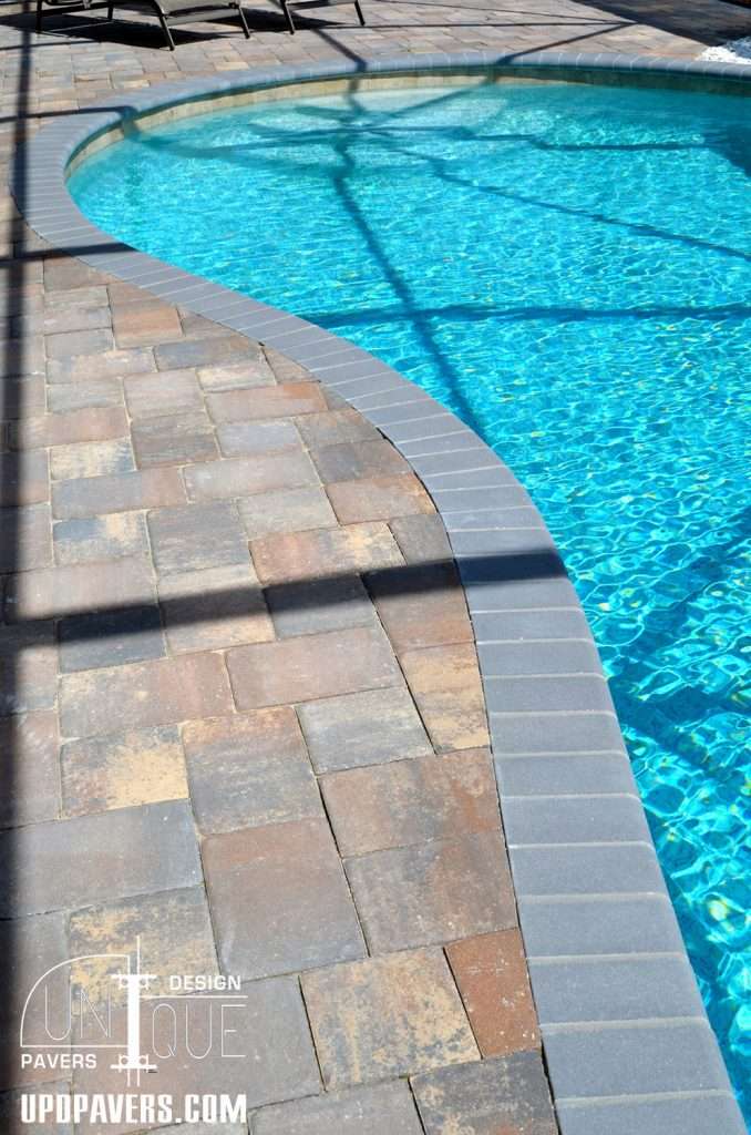 Curved paver pool deck with clear blue water reflecting sunlight. Unique design features.