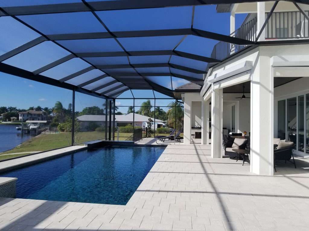 Modern outdoor pool with screened enclosure next to a waterfront home. Bright blue skies above.
