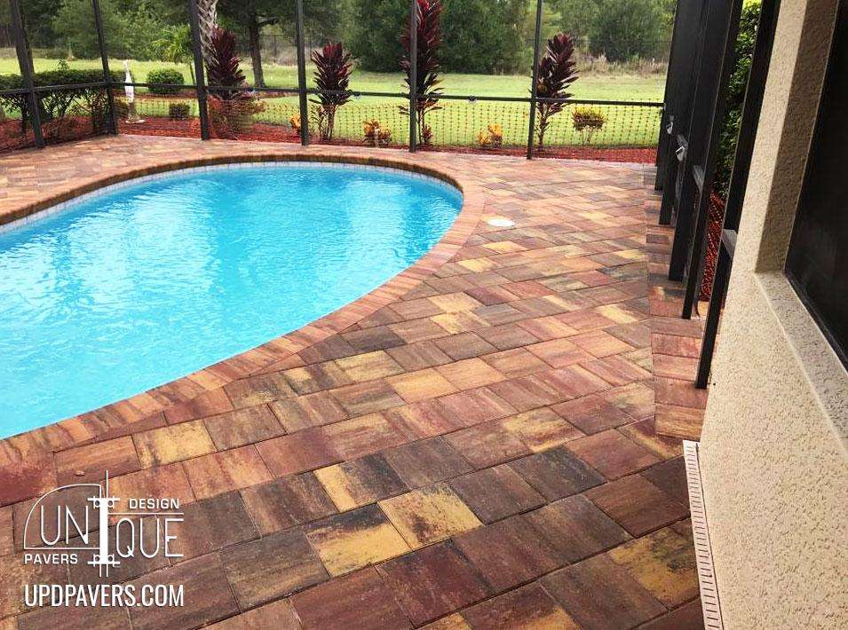 Poolside paved patio featuring colorful brick pavers and lush green landscaping.