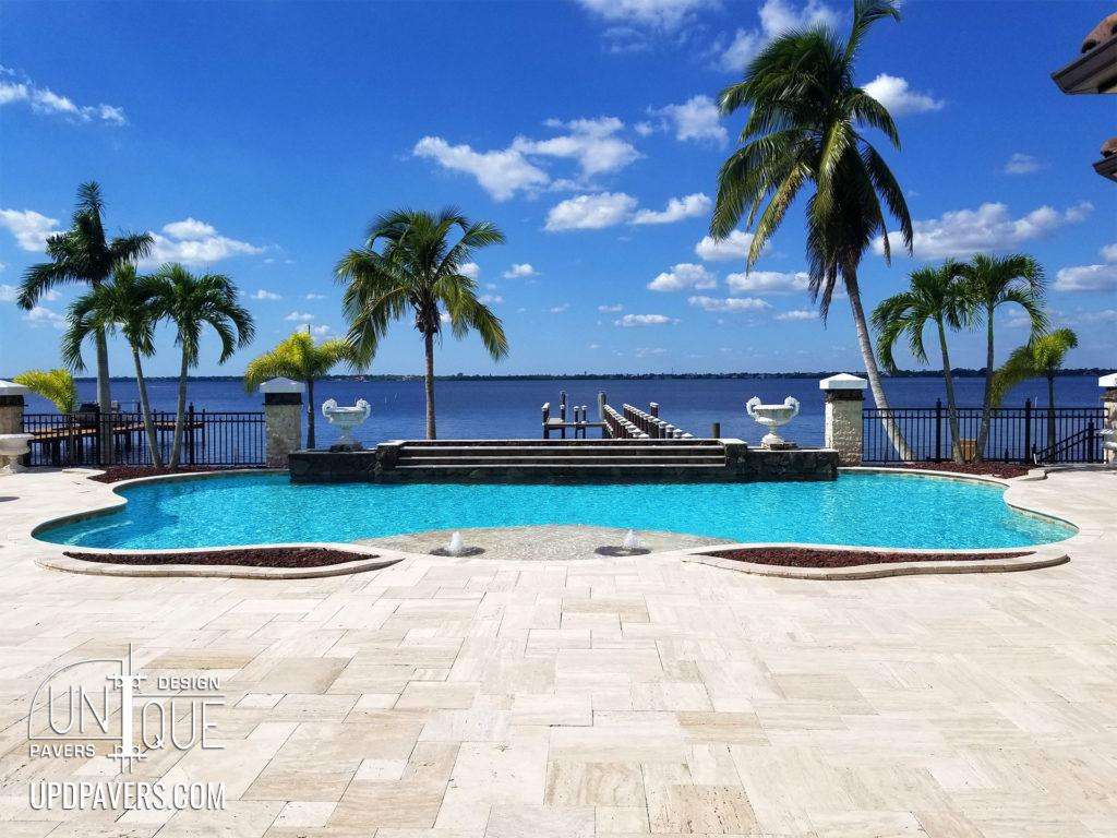 Luxury pool with pavers, palm trees, and waterfront view under a blue sky.