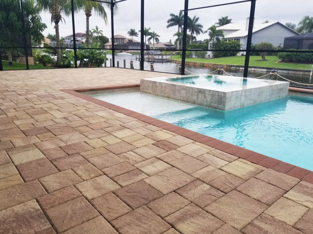 Paved patio with a pool and spa surrounded by palm trees, overlooking a waterfront view.