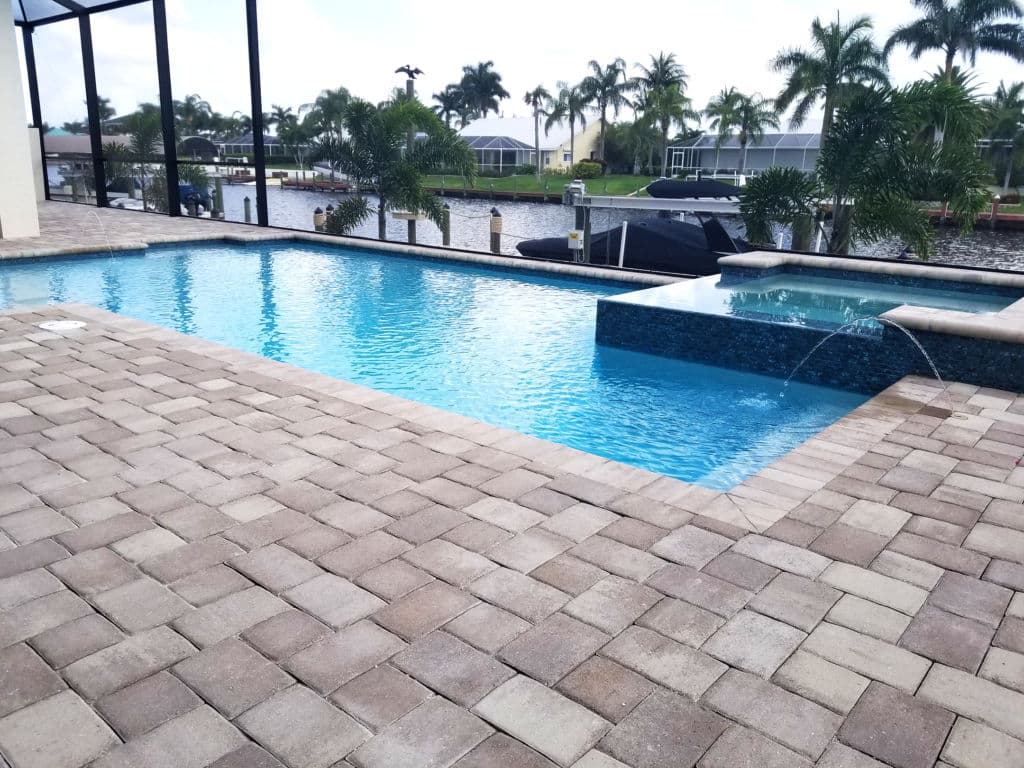 Modern backyard pool with spa, pavers, and waterfront view surrounded by palm trees.
