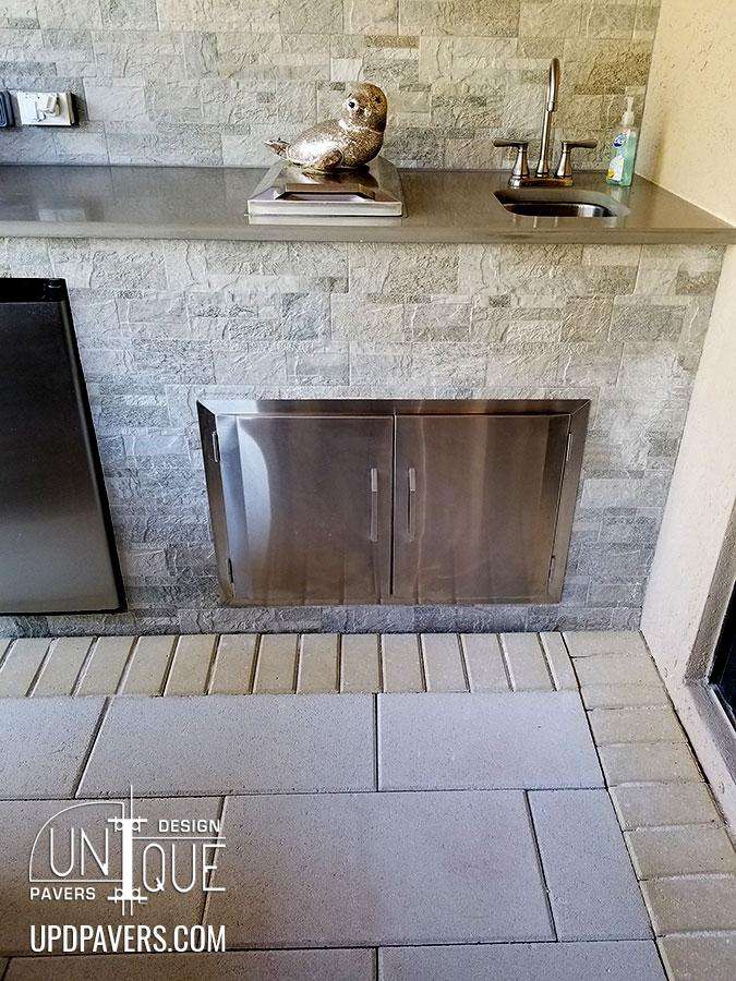 Modern outdoor kitchen with stainless steel storage, stone walls, and paver flooring.