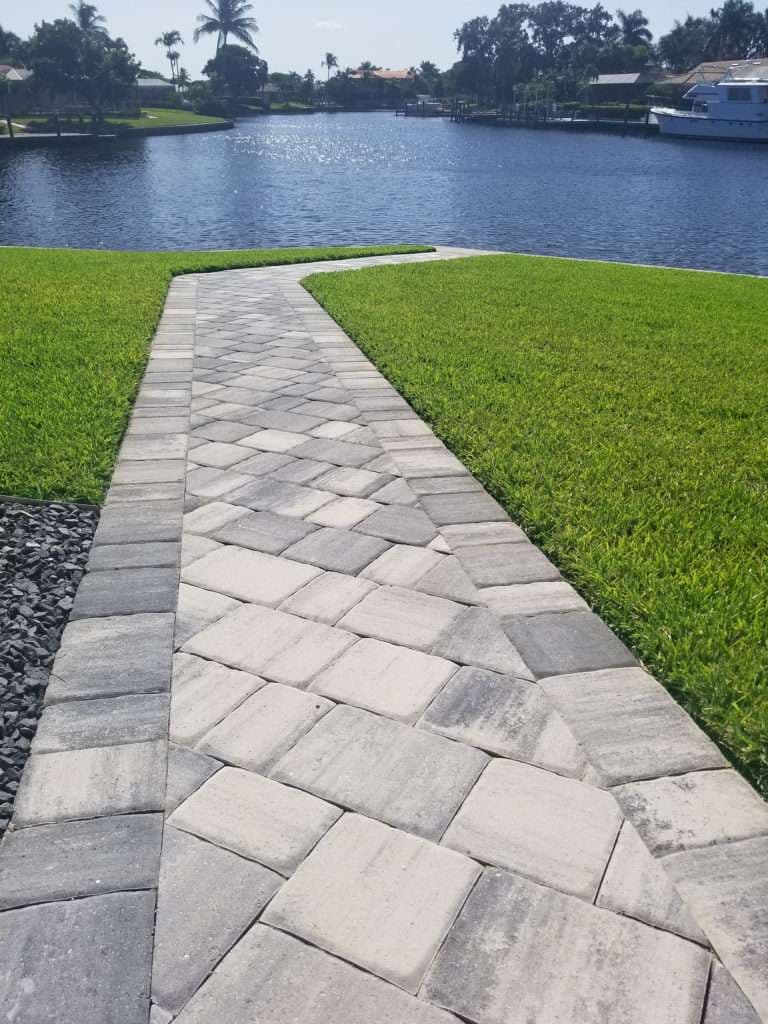 Paved walkway leading to a tranquil waterfront with lush green grass and palm trees.