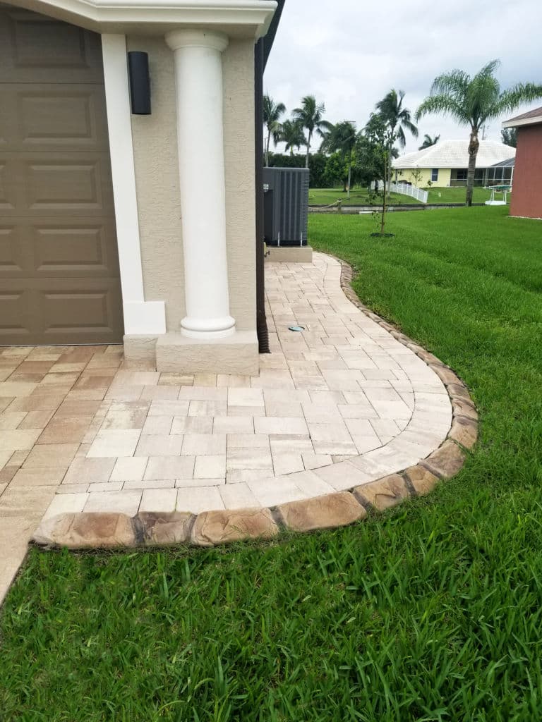 Paved walkway beside a house, lined with grass and tropical landscape features.