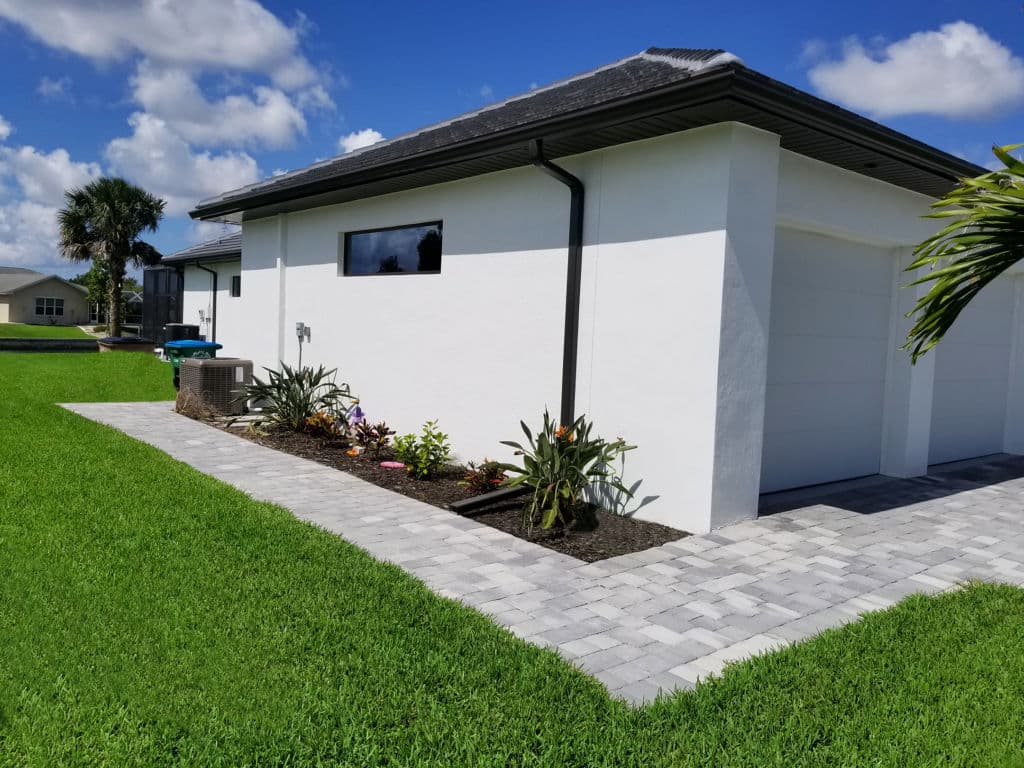 Modern white house exterior with landscaped garden and paved walkway under a blue sky.