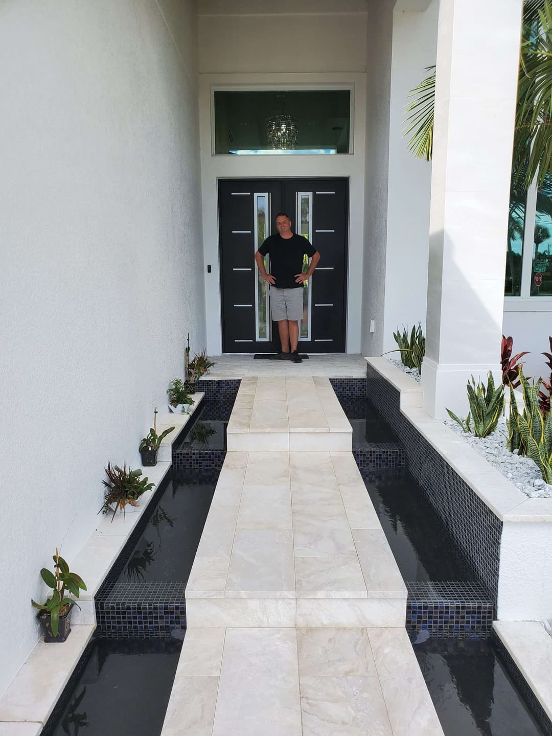 Man standing at a modern home entrance with a stone pathway and decorative plants.