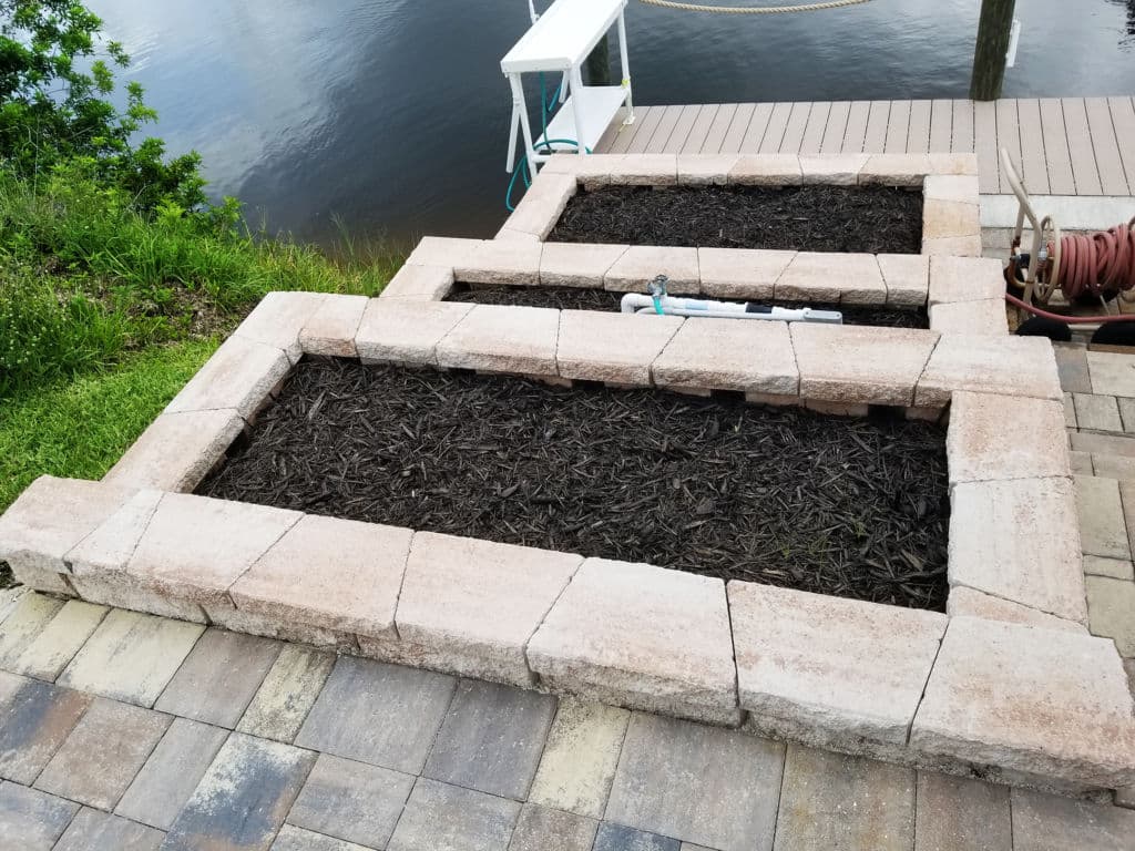 Raised garden beds made of stone blocks beside a calm water body, with mulch inside.