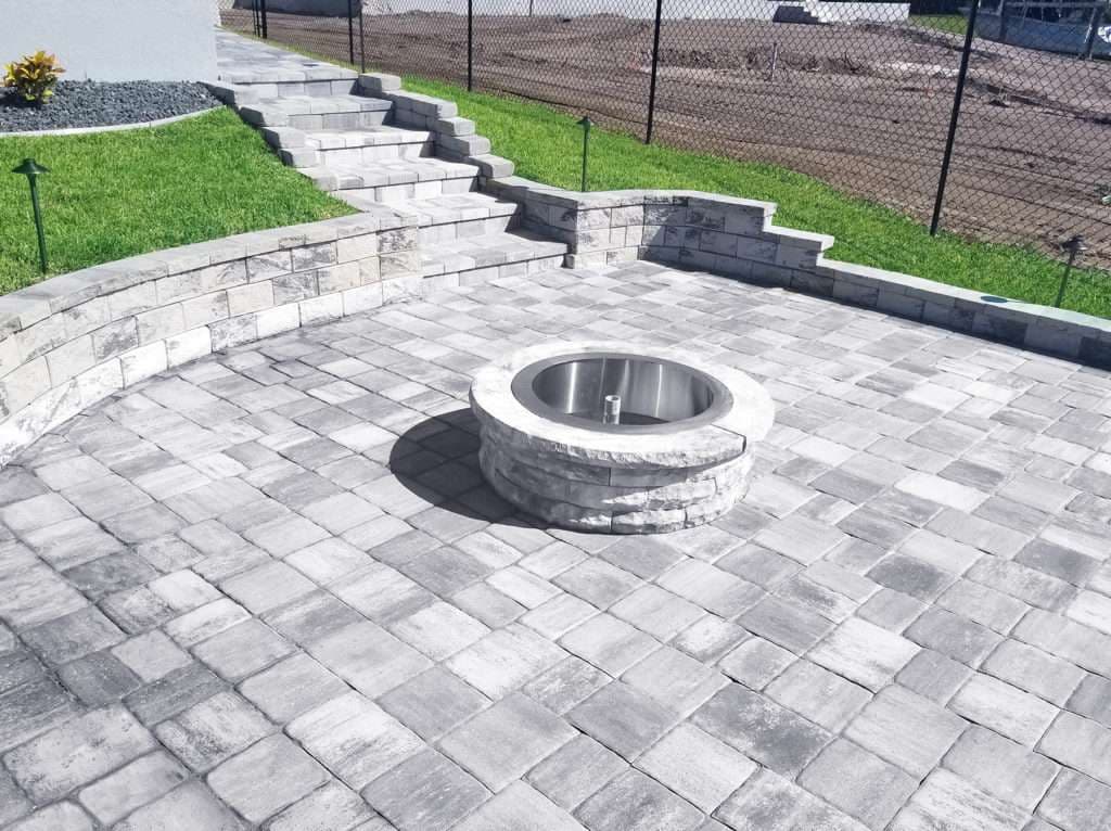 Outdoor patio with a circular stone fire pit and decorative pavers, surrounded by landscaped grass.