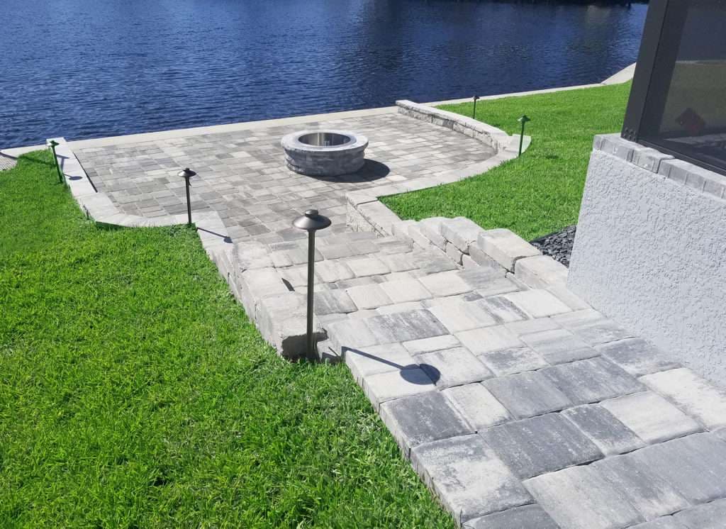 Paver patio with fire pit and pathway by a serene lake, surrounded by lush green grass.