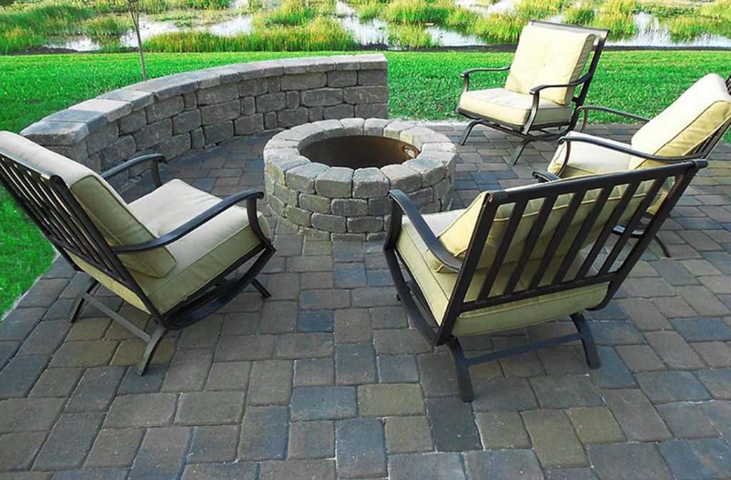 Outdoor patio with four chairs around a stone fire pit, surrounded by grass and water.