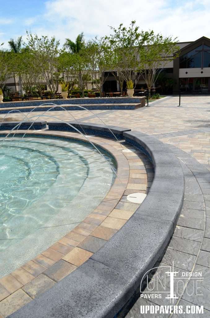 Modern outdoor pool area with fountains, paver stones, and lush landscaping.