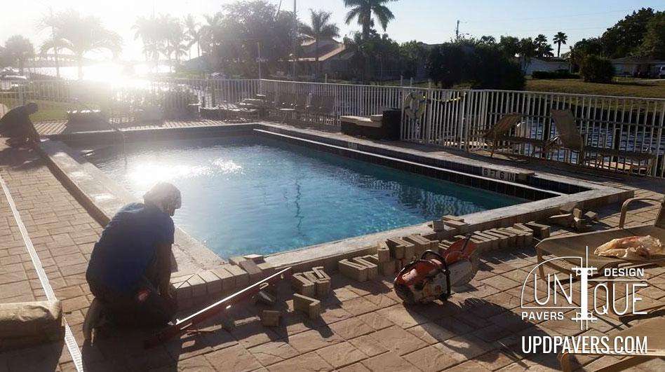 Worker installing pavers around a pool by the water with palm trees in the background.