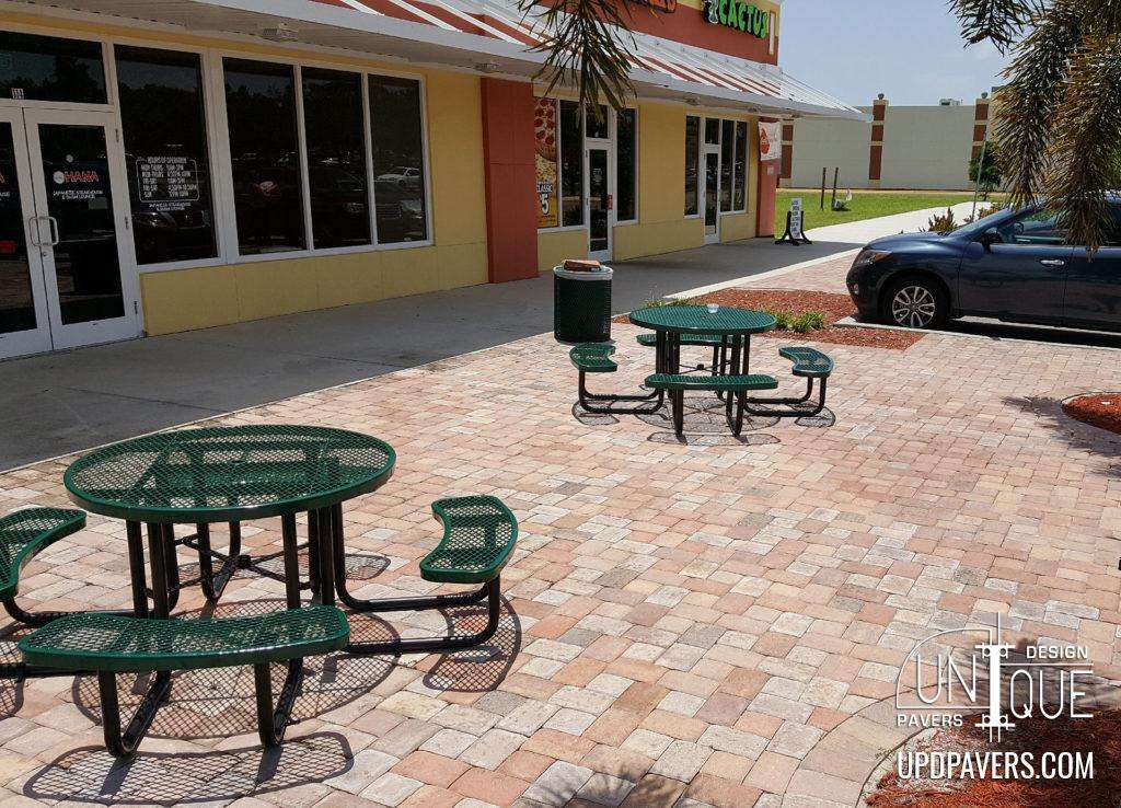 Green picnic tables on a paver patio outside a retail store, surrounded by landscaping.