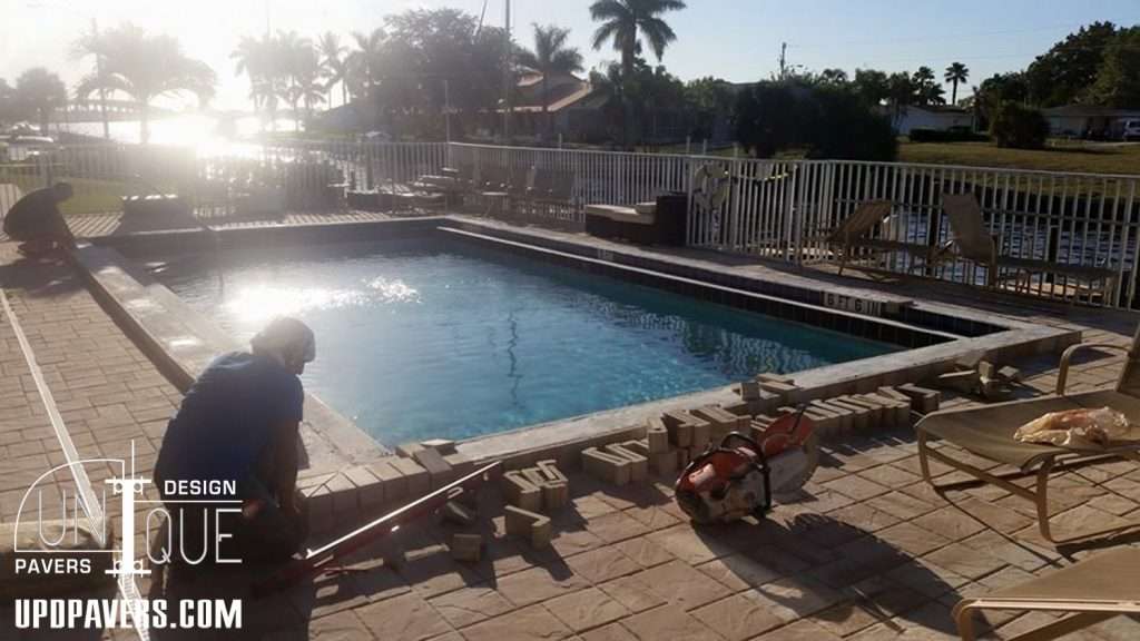 Pool renovation in progress with workers laying pavers under clear blue sky.