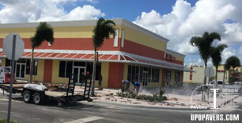Construction workers installing pavers outside a retail building under a blue sky.