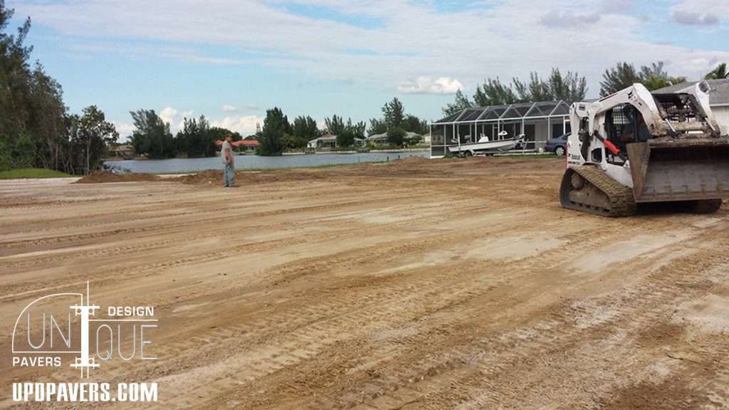Construction site with heavy machinery preparing land near a water body. Unique Pavers logo.