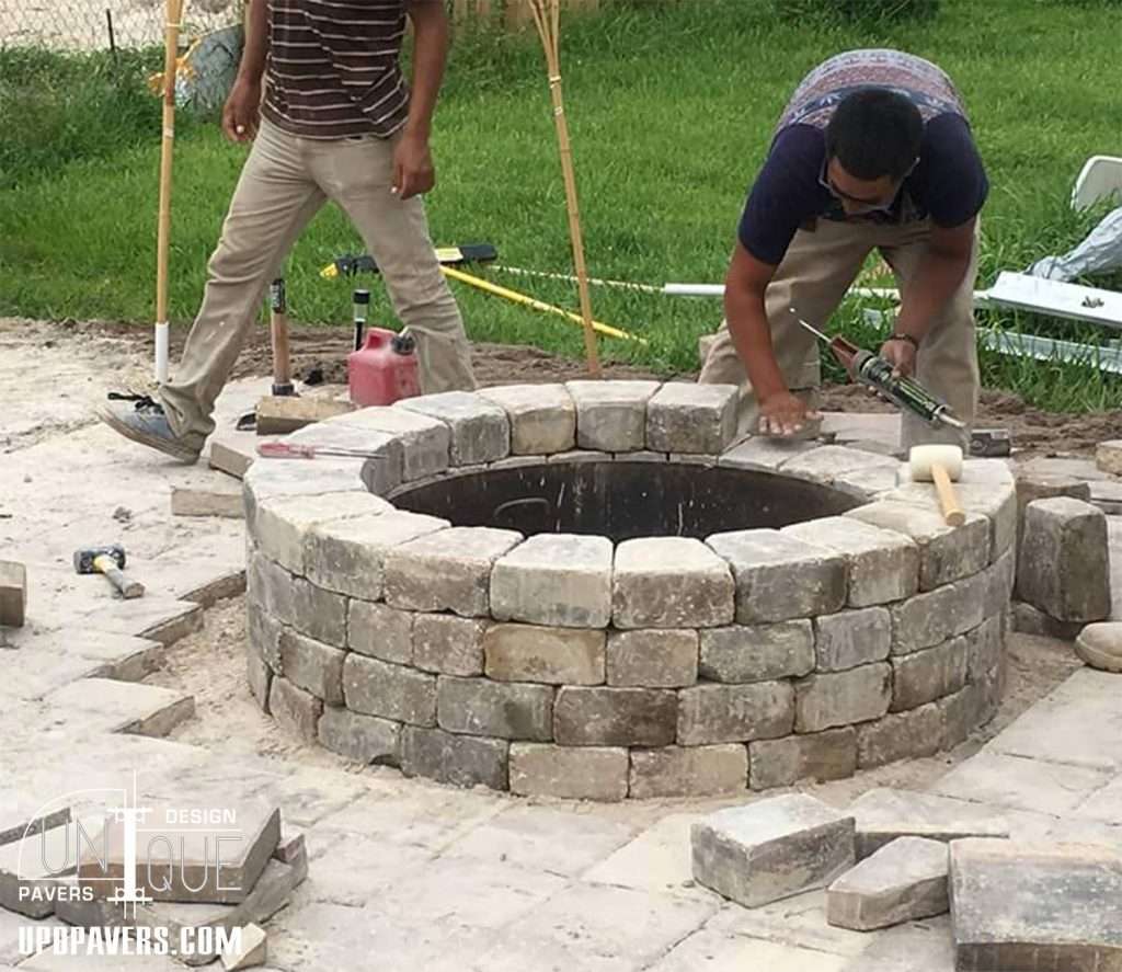 Two workers constructing a stone fire pit using pavers on a grass area.