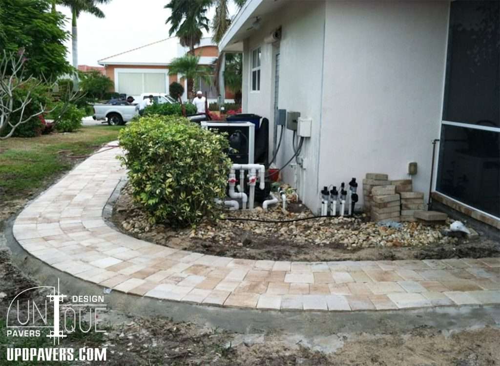 Paver walkway curving around a house with landscaping and utility features nearby.