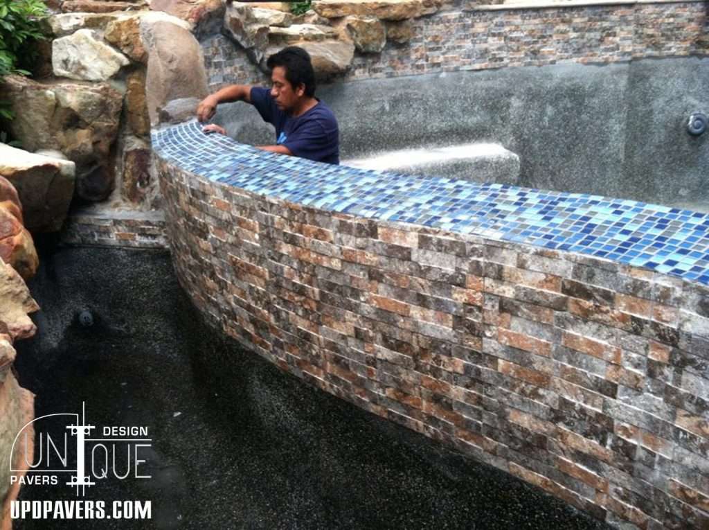 Mason installing mosaic tile on a curved stone wall in a spa area. Unique Pavers design.