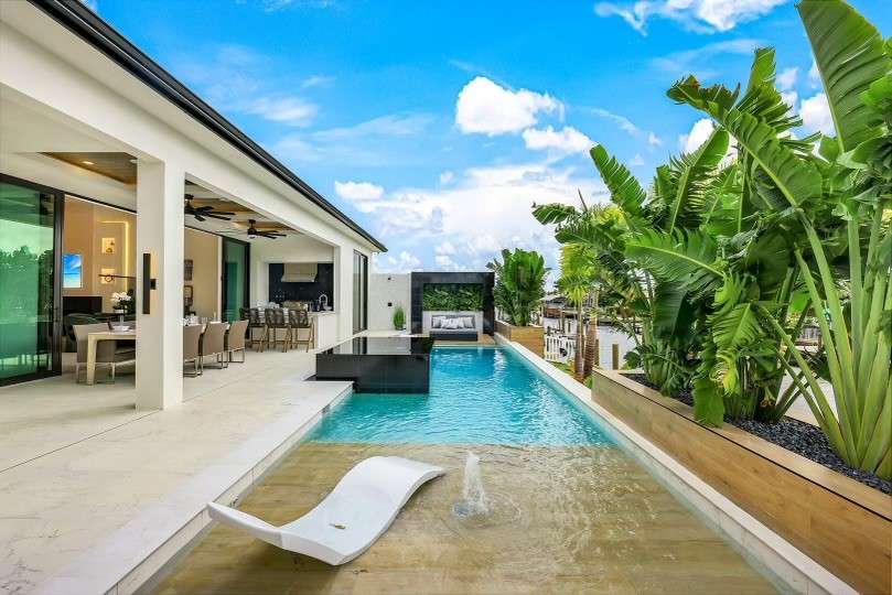 Modern luxury home with a pool, outdoor seating, and lush greenery under a bright blue sky.