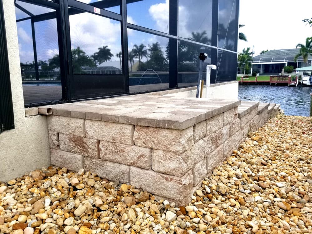Paver stone patio by water with river rock landscaping and screened enclosure in background.